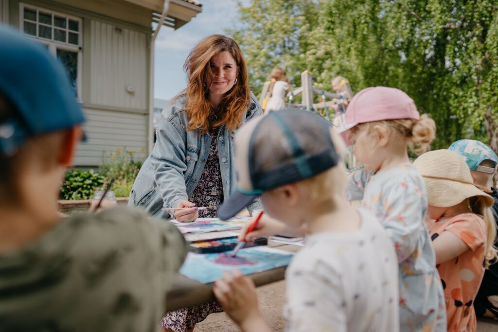 Aikuinen ohjaa pienten lasten maalaushetkeä ulkona pöydän äärellä