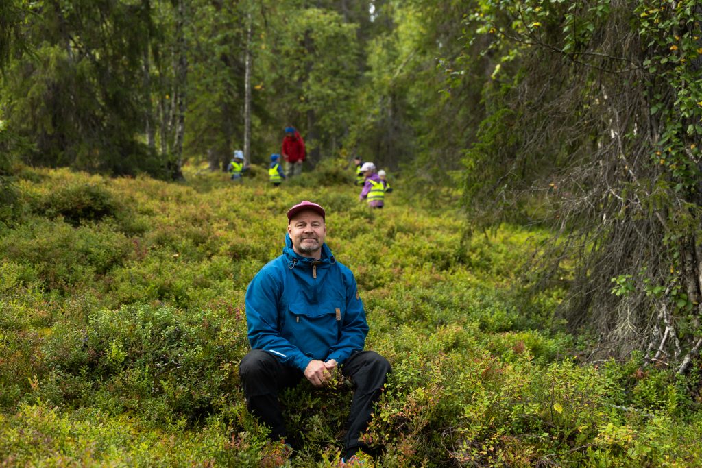 Janne Kylmämaa istuu kanervikossa, taustalla lapsiryhmä huomioliiveissä retkellä metsässä, Äkäslompolo