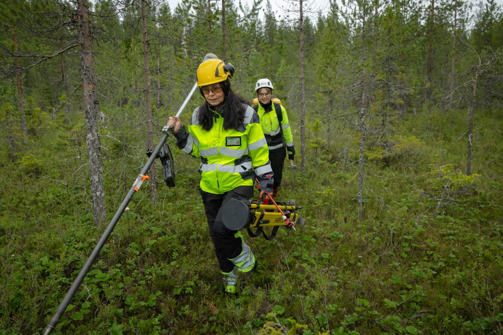 Merja Mustonen ja Mira Moberg kulkevat suojavaatteissa ja kypärissä metsässä mittalaitteiden kanssa, Rovaniemi